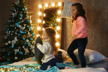 Little friendly sisters fight pillows on Christmas Eve. Children play and have fun together waiting for presents for the holiday. Room in bright christmas lights. Holiday decorations.