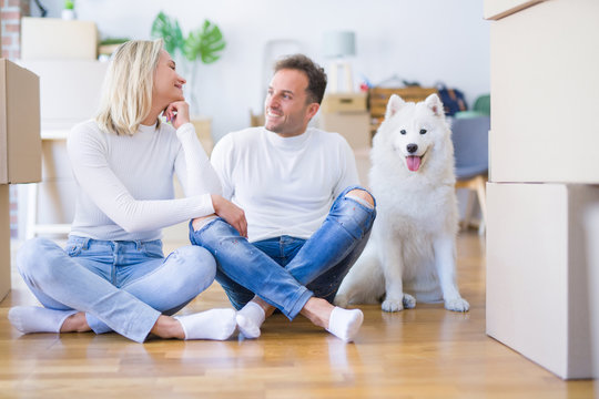 Young beautiful couple with dog sitting on the floor at new home around cardboard boxes