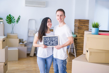Fototapeta na wymiar Young beautiful couple standing on the floor holding blackboard with message at new home around cardboard boxes