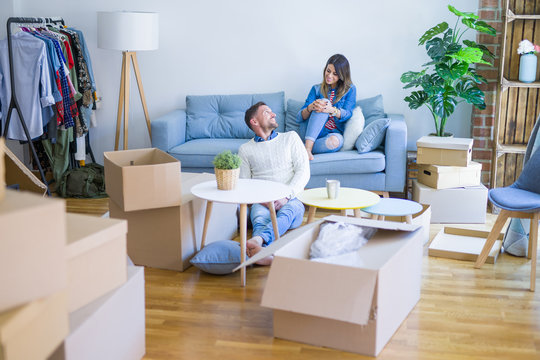 Young beautiful couple sitting on the sofa drinking cup of coffee at new home around cardboard boxes