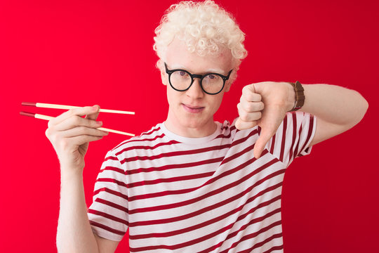 Young albino blond man holding chopsticks standing over isolated white background with angry face, negative sign showing dislike with thumbs down, rejection concept