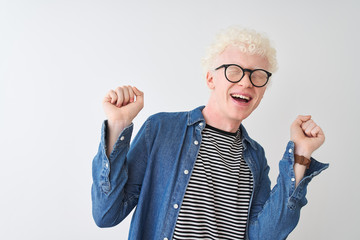 Young albino blond man wearing denim shirt and glasses over isolated white background celebrating surprised and amazed for success with arms raised and eyes closed. Winner concept.