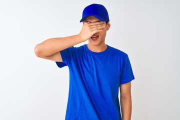 Chinese deliveryman wearing blue t-shirt and cap standing over isolated white background smiling and laughing with hand on face covering eyes for surprise. Blind concept.
