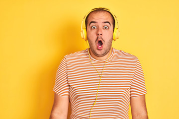 Young man listening to music using headphones standing over isolated yellow background afraid and shocked with surprise expression, fear and excited face.
