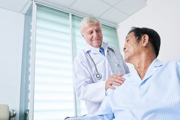Smiling general practitioner patting shoulder of senior Vietnamese man to support him during...