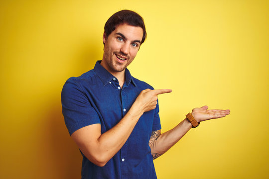 Young handsome man with tattoo wearing casual shirt standing over isolated yellow background amazed and smiling to the camera while presenting with hand and pointing with finger.