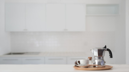 Coffee cup and Moka pot in wooden tray on white counter with blurred kitchen background