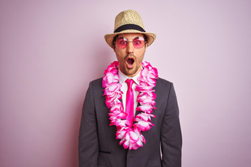 Young businessman wearing suit hat glasses hawaiian lei over isolated pink background scared in...