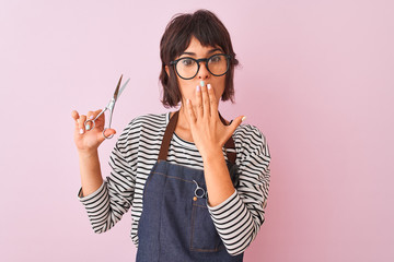 Hairdresser woman wearing apron and glasses holding scissors over isolated pink background cover mouth with hand shocked with shame for mistake, expression of fear, scared in silence, secret concept