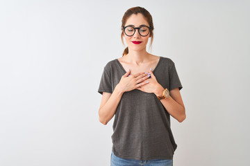 Redhead woman wearing casual t-shirt and glasses standing over isolated white background smiling with hands on chest with closed eyes and grateful gesture on face. Health concept.