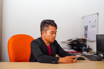Young Asian man sitting in office doing work on phone or desktop computer in casual outfit.