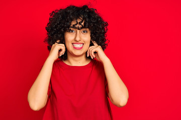 Young arab woman with curly hair wearing casual t-shirt over isolated red background covering ears with fingers with annoyed expression for the noise of loud music. Deaf concept.