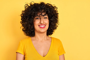 Young arab woman with curly hair wearing t-shirt standing over isolated yellow background with a happy face standing and smiling with a confident smile showing teeth