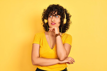 Arab woman with curly hair listening to music using headphones over isolated yellow background with hand on chin thinking about question, pensive expression. Smiling with thoughtful face. Doubt