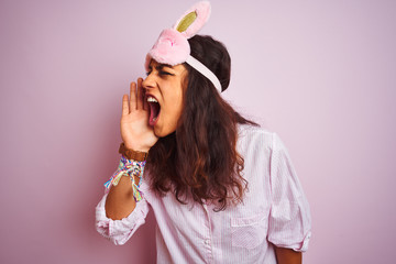 Young beautiful woman wearing pajama and sleep mask over isolated pink background shouting and screaming loud to side with hand on mouth. Communication concept.