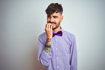 Young man with tattoo wearing purple shirt and bow tie over isolated white background looking stressed and nervous with hands on mouth biting nails. Anxiety problem.