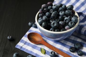 Fresh ripe blueberries are poured into a ceramic blue bowl on a checkered towel and a background of dark wood and a brown wooden spoon. Healthy snack concept. Useful antioxidants.