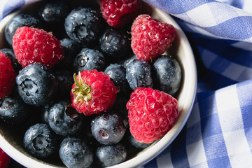 Fresh ripe blueberries and raspberries are in a ceramic bowl against a checkered white and blue towel. Berries close-up. Healthy organic food concept. Useful snack. Antioxidant diet.