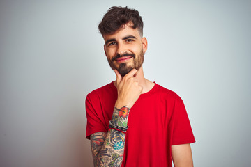 Young man with tattoo wearing red t-shirt standing over isolated white background looking confident at the camera smiling with crossed arms and hand raised on chin. Thinking positive.