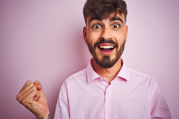 Young man with tattoo wearing shirt standing over isolated pink background celebrating surprised and amazed for success with arms raised and open eyes. Winner concept.