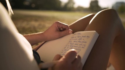 Close up on a diary while a young lady writes in it at a golden sunset in a park. She wears shorts and the sun is shining on her skin