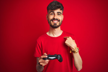 Fototapeta na wymiar Young gamer man with tattoo playing video game standing over isolated red background with surprise face pointing finger to himself
