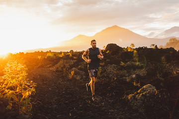 Young male athlete trail running in mountains at sunrise. Amazing black lava volcanic landscape of...