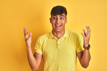 Young indian man wearing casual polo standing over isolated yellow background celebrating crazy and amazed for success with arms raised and open eyes screaming excited. Winner concept