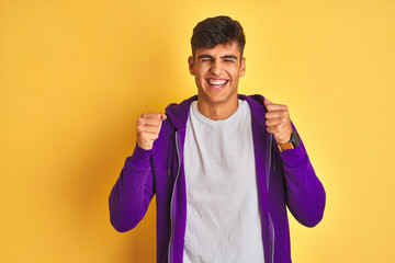 Young indian man wearing purple sweatshirt standing over isolated yellow background excited for success with arms raised and eyes closed celebrating victory smiling. Winner concept.