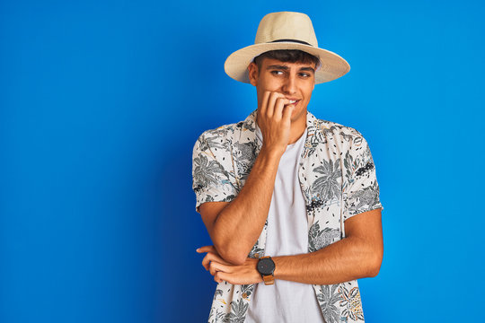 Indian man on vacation wearing hawiaian shirt summer hat over isolated blue background looking stressed and nervous with hands on mouth biting nails. Anxiety problem.