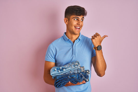 Young indian shopkeeper man holding folded jeans standing over isolated pink background pointing and showing with thumb up to the side with happy face smiling