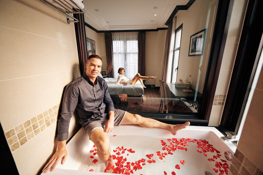 Smiling young man preparing romantic hot bath with rose petals for him and his beautiful girlfriend