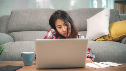 Freelance Asian woman working at home, business female working on laptop and using mobile phone talking with customer on sofa in living room at home. Lifestyle women working at home concept.