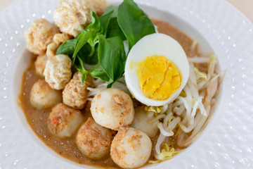 Thai Food Rice Noodles in Fish Curry With boiled egg, fish balls, pork snack, basil, bean sprouts on top, placed on a wooden table.