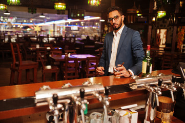 Handsome well-dressed arabian man with glass of whiskey and cigar posed at pub.