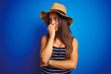 Young beautiful woman wearing striped t-shirt and summer hat over isolated blue background looking stressed and nervous with hands on mouth biting nails. Anxiety problem.