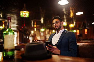 Handsome well-dressed arabian man with glass of whiskey and cigar hold mobile phone,  posed at pub.