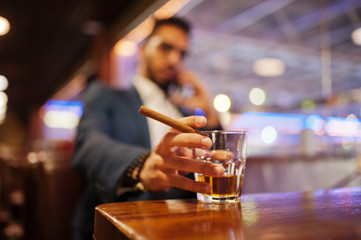 Close up hand of handsome well-dressed arabian man with glass of whiskey and cigar posed at pub.