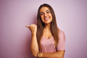 Young beautiful woman wearing t-shirt standing over isolated pink background smiling with happy face looking and pointing to the side with thumb up.