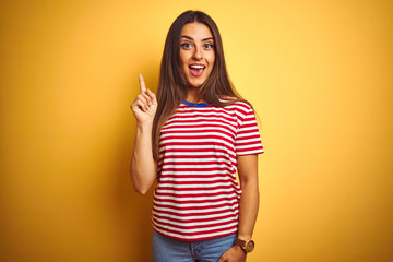 Young beautiful woman wearing striped t-shirt standing over isolated yellow background pointing finger up with successful idea. Exited and happy. Number one.