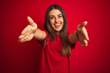 Young beautiful woman wearing t-shirt standing over isolated red background looking at the camera smiling with open arms for hug. Cheerful expression embracing happiness. - obrazy, fototapety, plakaty