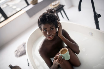Dark-eyed woman drinking morning coffee while having bath