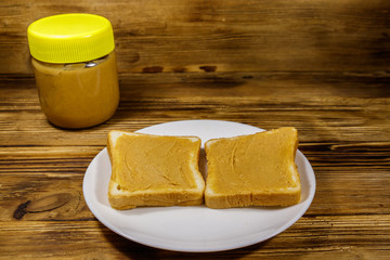 Jar of peanut butter and plate with two sandwiches with peanut butter on wooden table