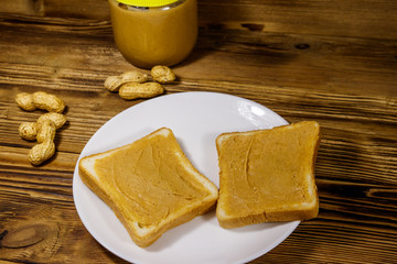 Jar of peanut butter and plate with two sandwiches with peanut butter on wooden table