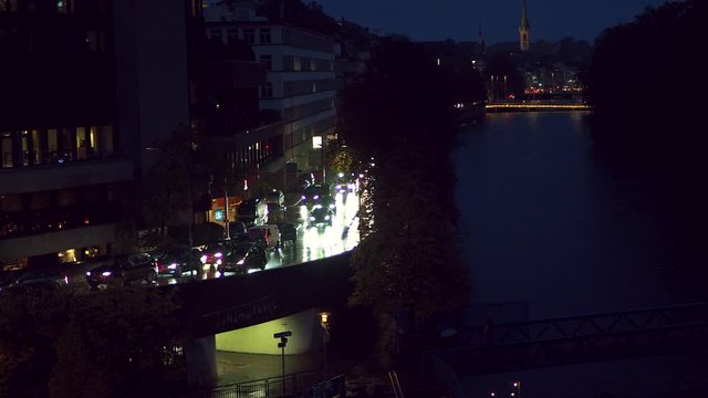 Cars driving down a wet road on a dark, rainy evening filmed in downtown Zurich, Switzerland along the Limmat River, church and trains in the background - Static Shot