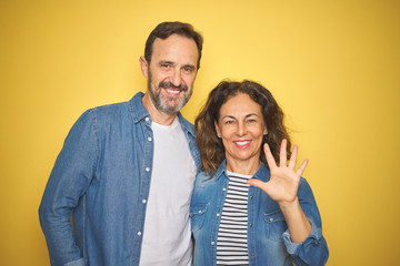 Beautiful middle age couple together wearing denim shirt over isolated yellow background showing and pointing up with fingers number five while smiling confident and happy.