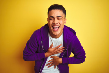 Young brazilian man wearing purple sweatshirt standing over isolated yellow background smiling and laughing hard out loud because funny crazy joke with hands on body.