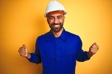 Handsome indian worker man wearing uniform and helmet over isolated yellow background celebrating...