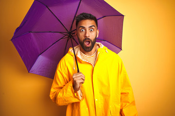 Young indian man wearing raincoat and purple umbrella over isolated yellow background scared in shock with a surprise face, afraid and excited with fear expression