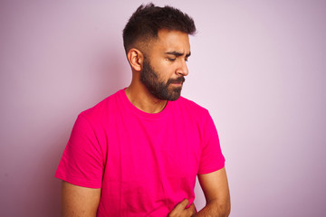 Young indian man wearing t-shirt standing over isolated pink background with hand on stomach because nausea, painful disease feeling unwell. Ache concept.